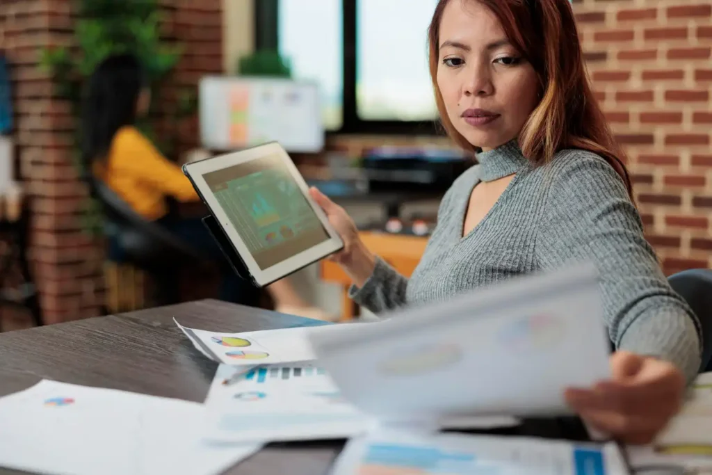 Mulher de cabelo vermelho analisando dados em tablet e documentos em ambiente de escritório, ilustrando o uso de Marketing Preditivo para avançar no marketing de dados
